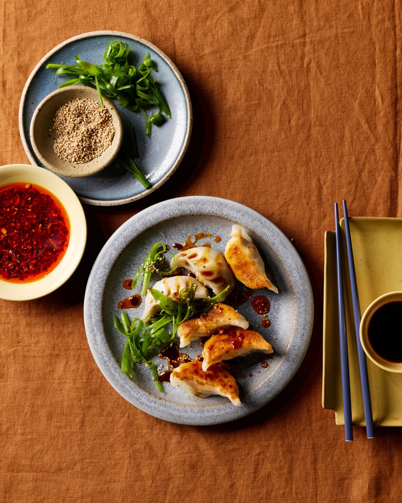 Pan-fried dumplings served on a round gray plate with green onions and chili oil, arranged on a textured brown tablecloth with dipping sauces and chopsticks.